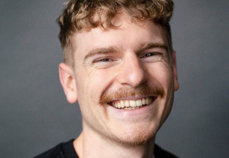 Paul Donati's headshot, showing him smiling into the camera in front of a grey background. He has chort, curly copperish hair and a moustache